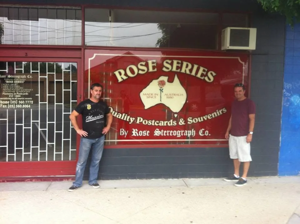Stephen and his brother Jeffery Cutts standing either side of the red Rose Series shopfront sign reading Quality Postcards and Souvenirs by Rose Stereograph Co., with an Australia map and rose emblem marked Made in Australia since 1880
