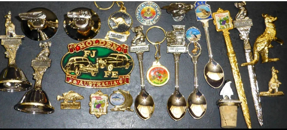 Sundowner Souvenirs products: gold and silver souvenir teaspoons, keyrings, bells, letter openers, badges, kangaroo and koala figures, a penguin bottle stopper and a Holden FJ FE Australia belt buckle